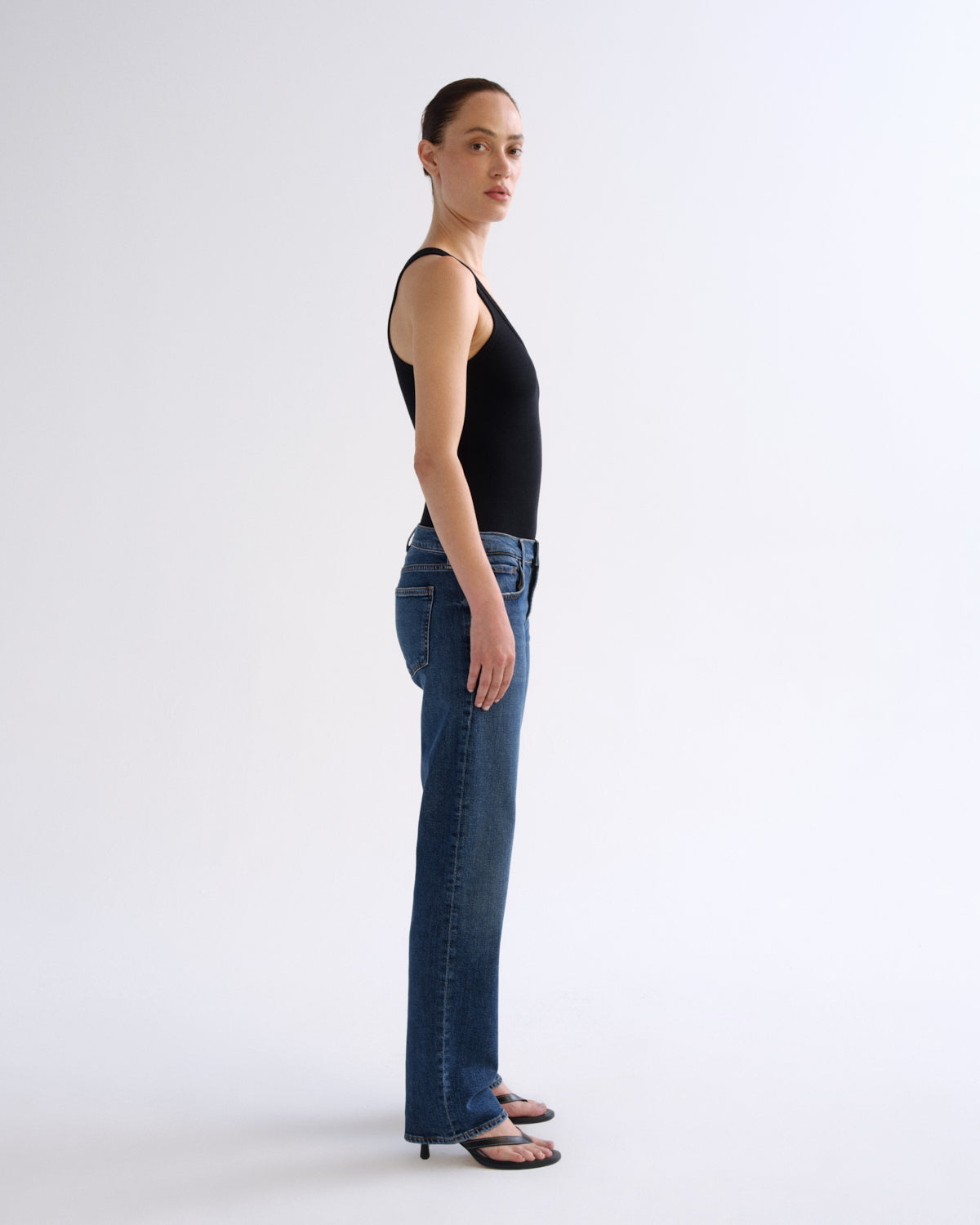 A woman stands in profile wearing a black sleeveless top, blue flare jeans, and black sandals against a clean white studio background.