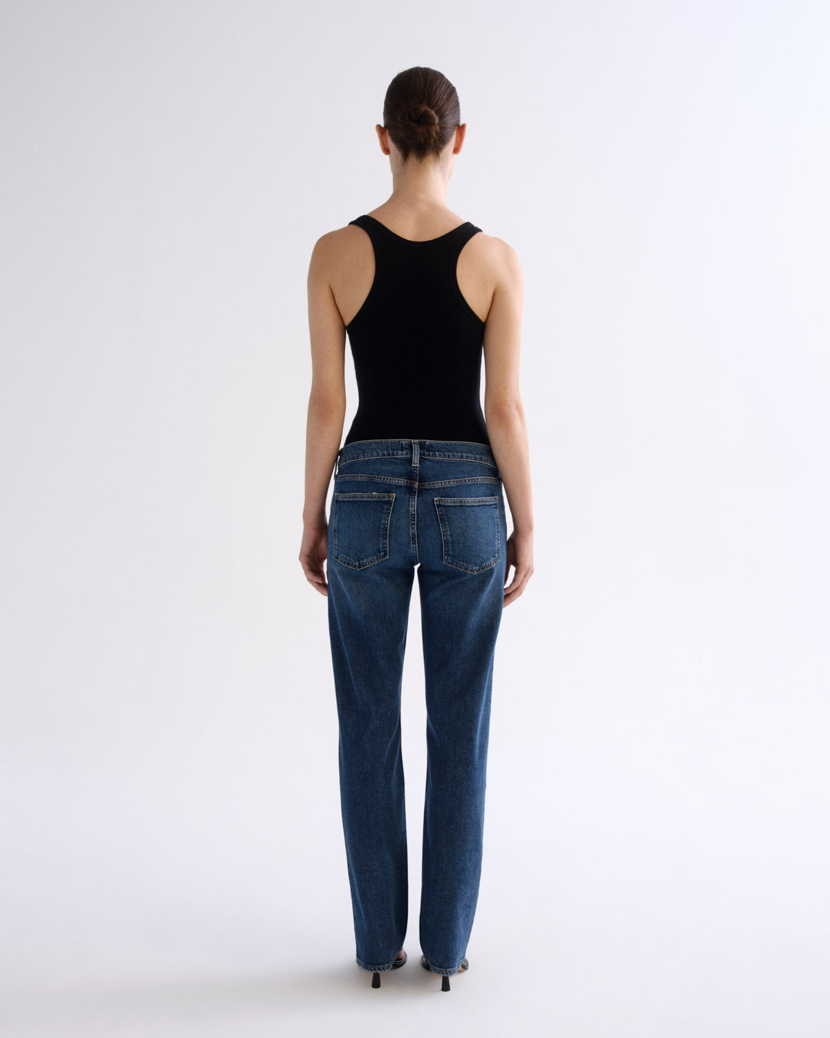 A woman viewed from behind wearing a black sleeveless top and blue jeans against a plain light gray studio background.