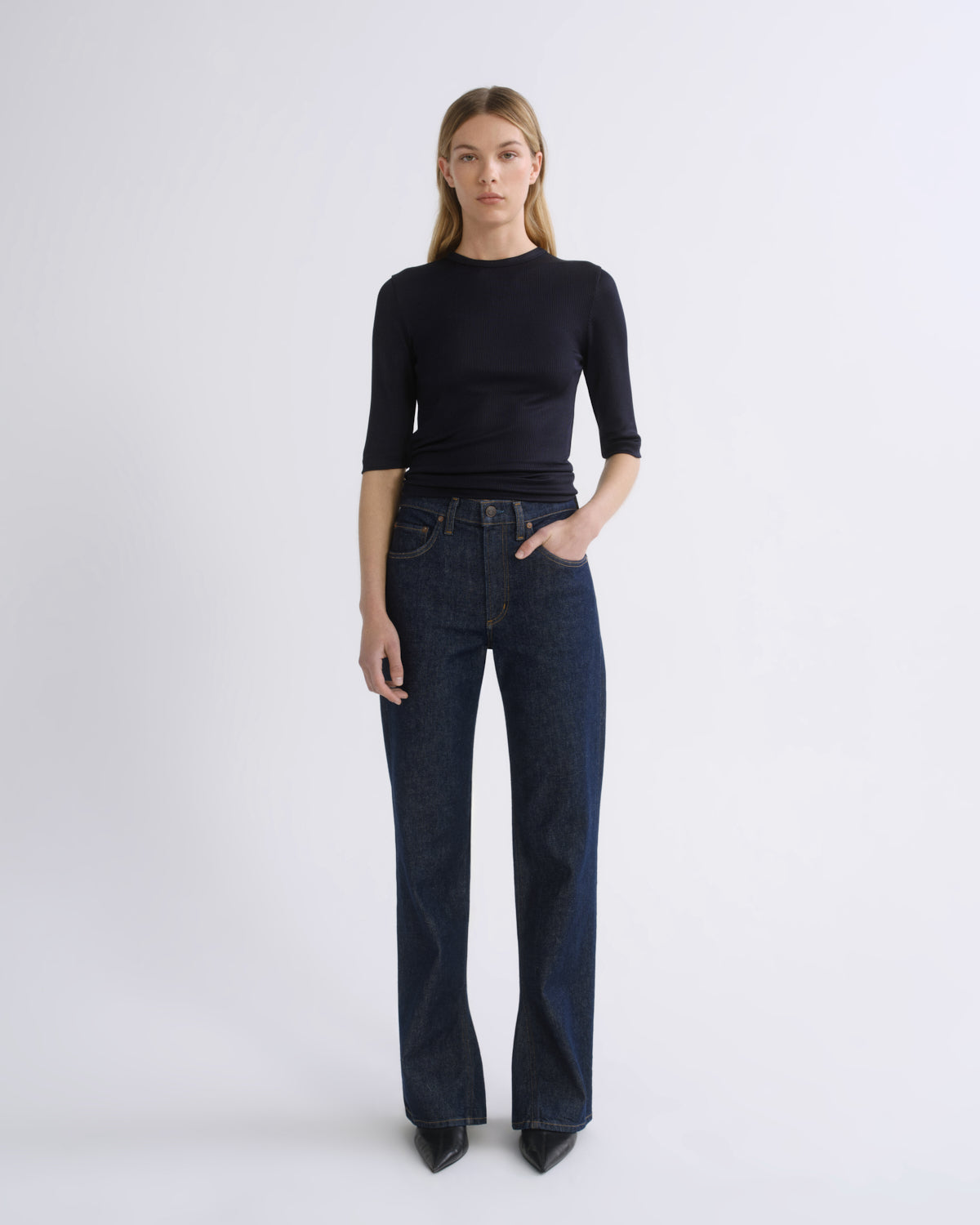 A woman stands against a light gray studio background wearing a fitted black three-quarter-sleeve top, dark blue straight-leg jeans, and black pointed shoes.