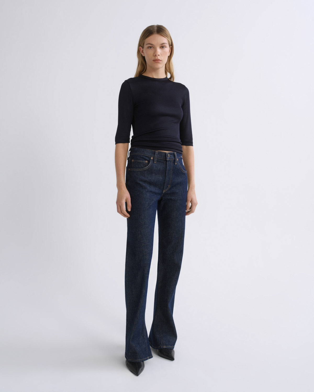 A model stands against a light gray studio background wearing a fitted black three-quarter-sleeve top and dark blue straight-leg jeans with black pointed shoes.