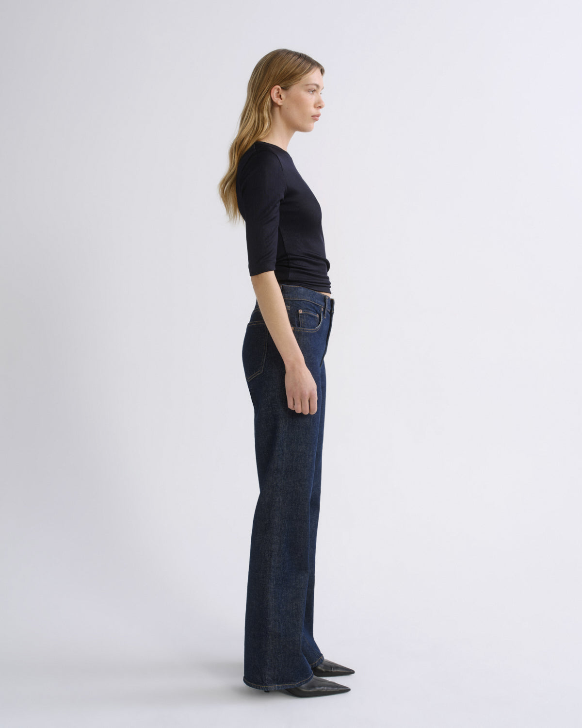 A woman stands in profile wearing a fitted black short-sleeve top and dark blue wide-leg jeans against a plain light gray studio background.