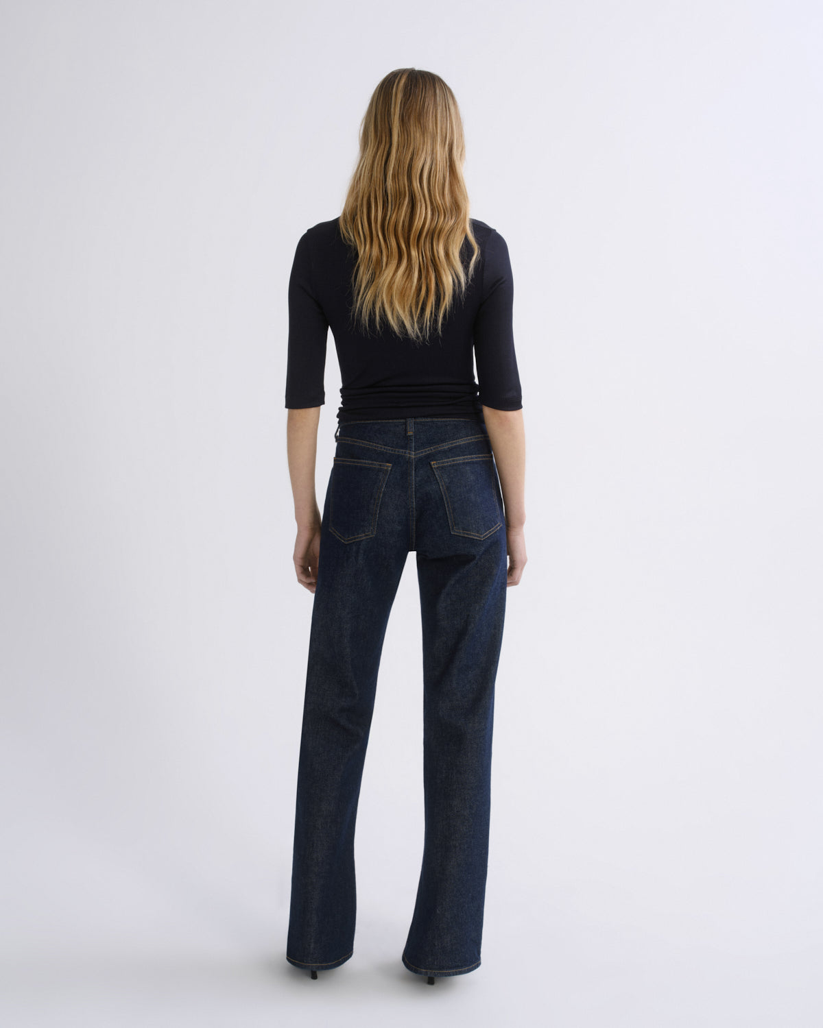 A woman viewed from the back wearing a black three-quarter sleeve top and dark blue straight-leg jeans against a light gray studio background.