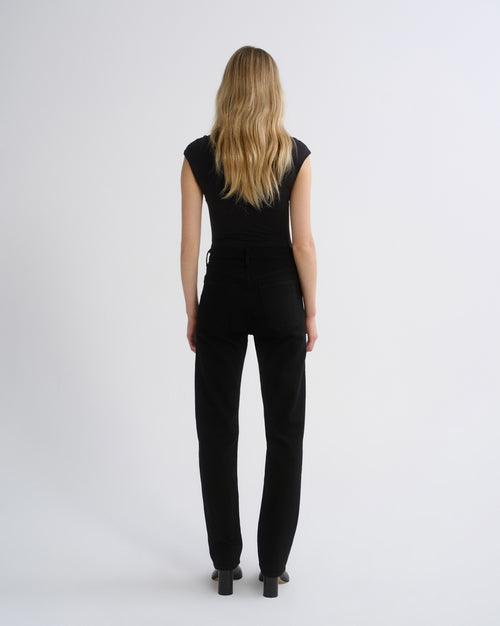 A woman with long blonde hair is shown from the back, wearing a black sleeveless top and black pants against a plain light gray studio background.