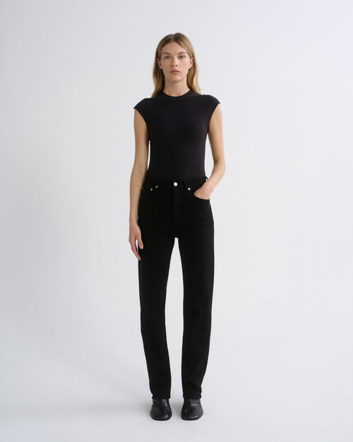 A woman modeling a black sleeveless mock-neck top and black straight-leg jeans against a plain light gray studio background.