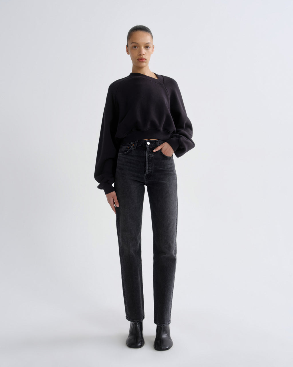 A model stands against a light gray studio background wearing a black long-sleeve crewneck sweatshirt, black high-waisted straight-leg jeans, and black ankle boots.