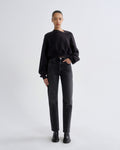 A model stands against a light gray studio background wearing a black long-sleeve crewneck sweatshirt, black high-waisted straight-leg jeans, and black ankle boots.