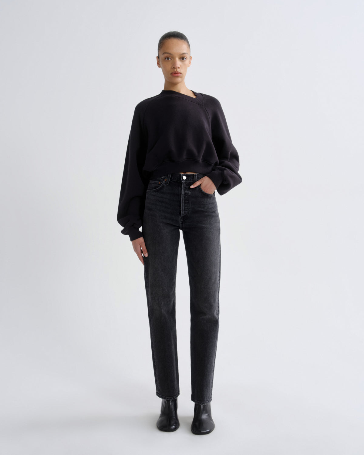 A model stands against a light gray studio background wearing a black long-sleeve crewneck sweatshirt, black high-waisted straight-leg jeans, and black ankle boots.