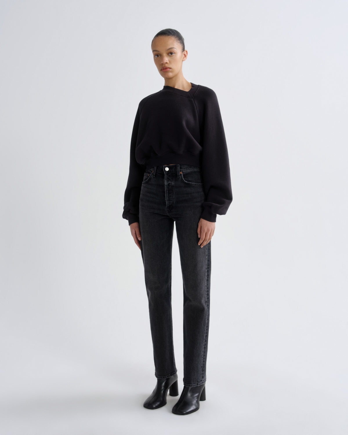A model stands against a light gray studio background wearing a black long-sleeve crewneck top, dark gray high-waisted straight-leg jeans, and black leather ankle boots.