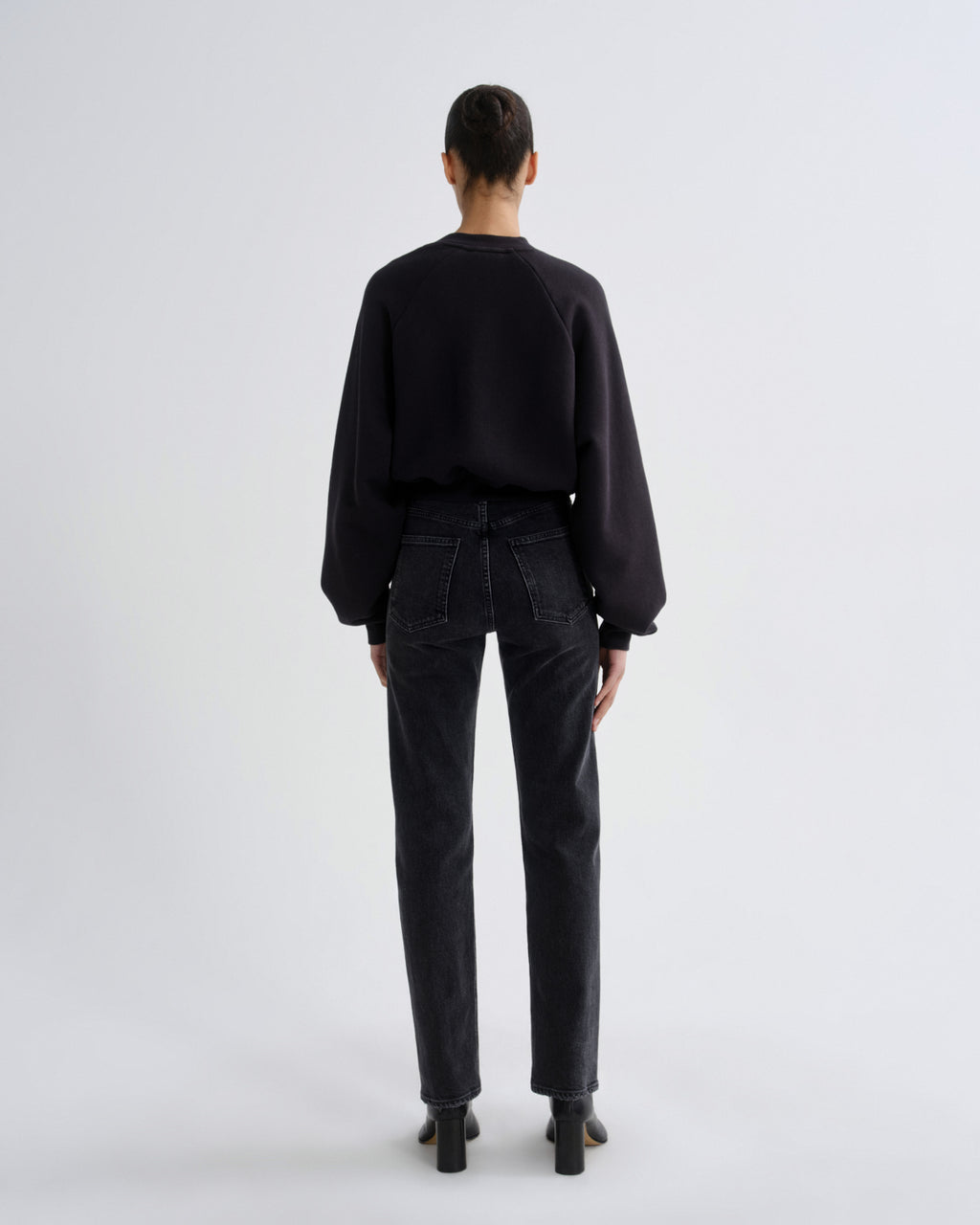 A model viewed from the back wearing a black long-sleeve crewneck sweatshirt, dark charcoal gray straight-leg jeans, and black block-heeled boots, set against a plain light gray studio background.