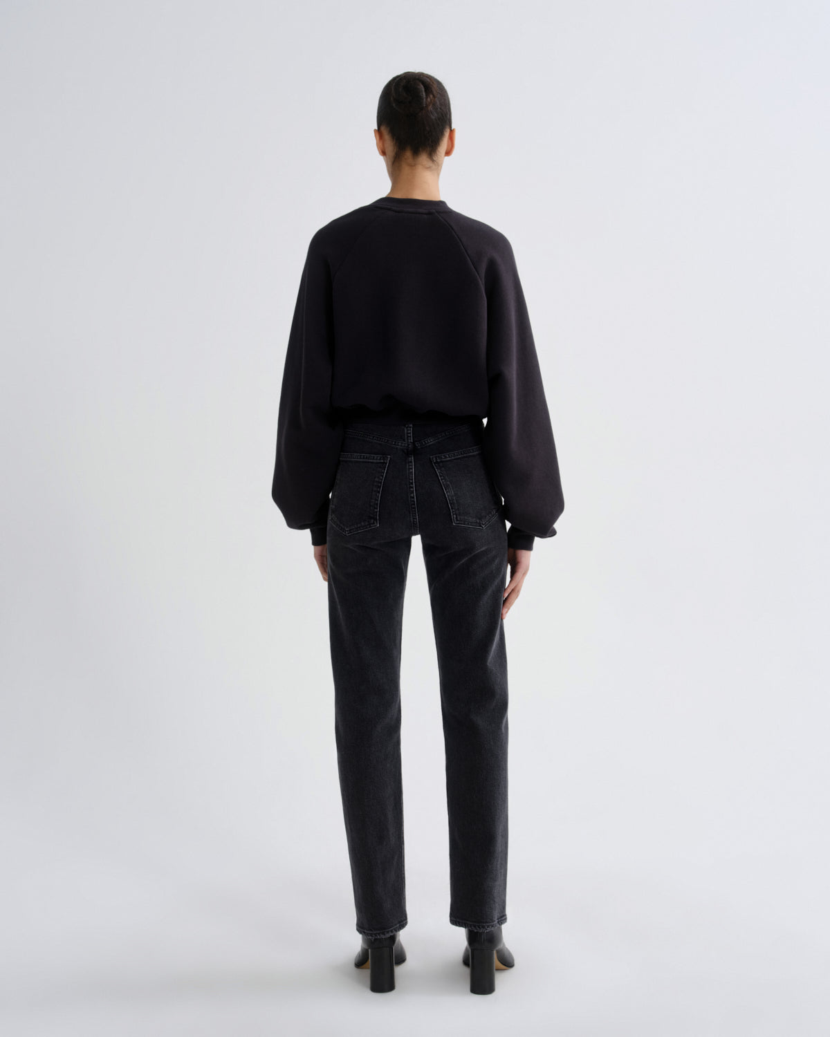 A model viewed from the back wearing a black long-sleeve crewneck sweatshirt, dark charcoal gray straight-leg jeans, and black block-heeled boots, set against a plain light gray studio background.