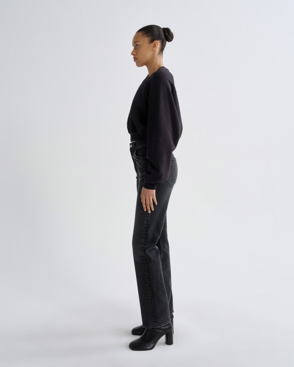 A model in profile wearing a black long-sleeve top, dark gray straight-leg jeans, and black heeled ankle boots, standing against a light gray studio background.