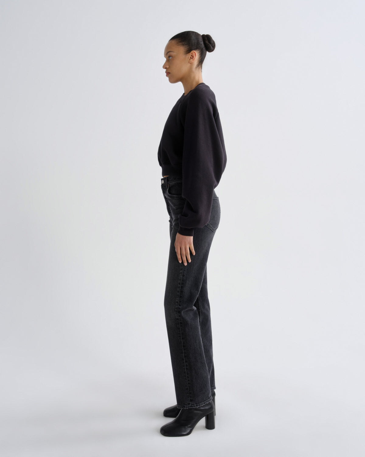 A model in profile wearing a black long-sleeve top, dark gray straight-leg jeans, and black heeled ankle boots, standing against a light gray studio background.