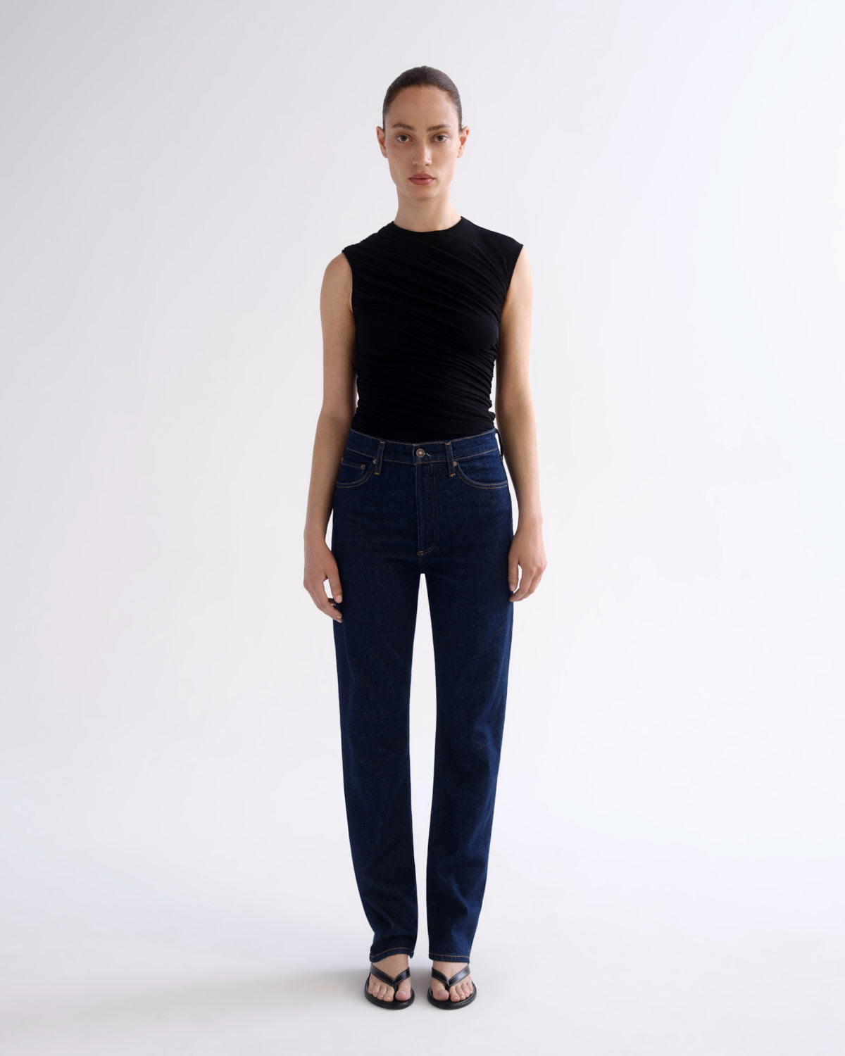 A model stands against a light gray studio background wearing a sleeveless black top and dark blue straight-leg jeans with black sandals.