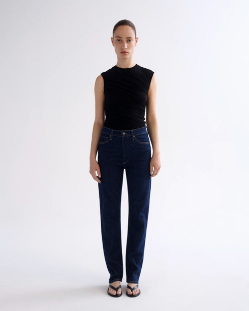 A model stands against a light gray studio background wearing a sleeveless black top and dark blue straight-leg jeans with black sandals.