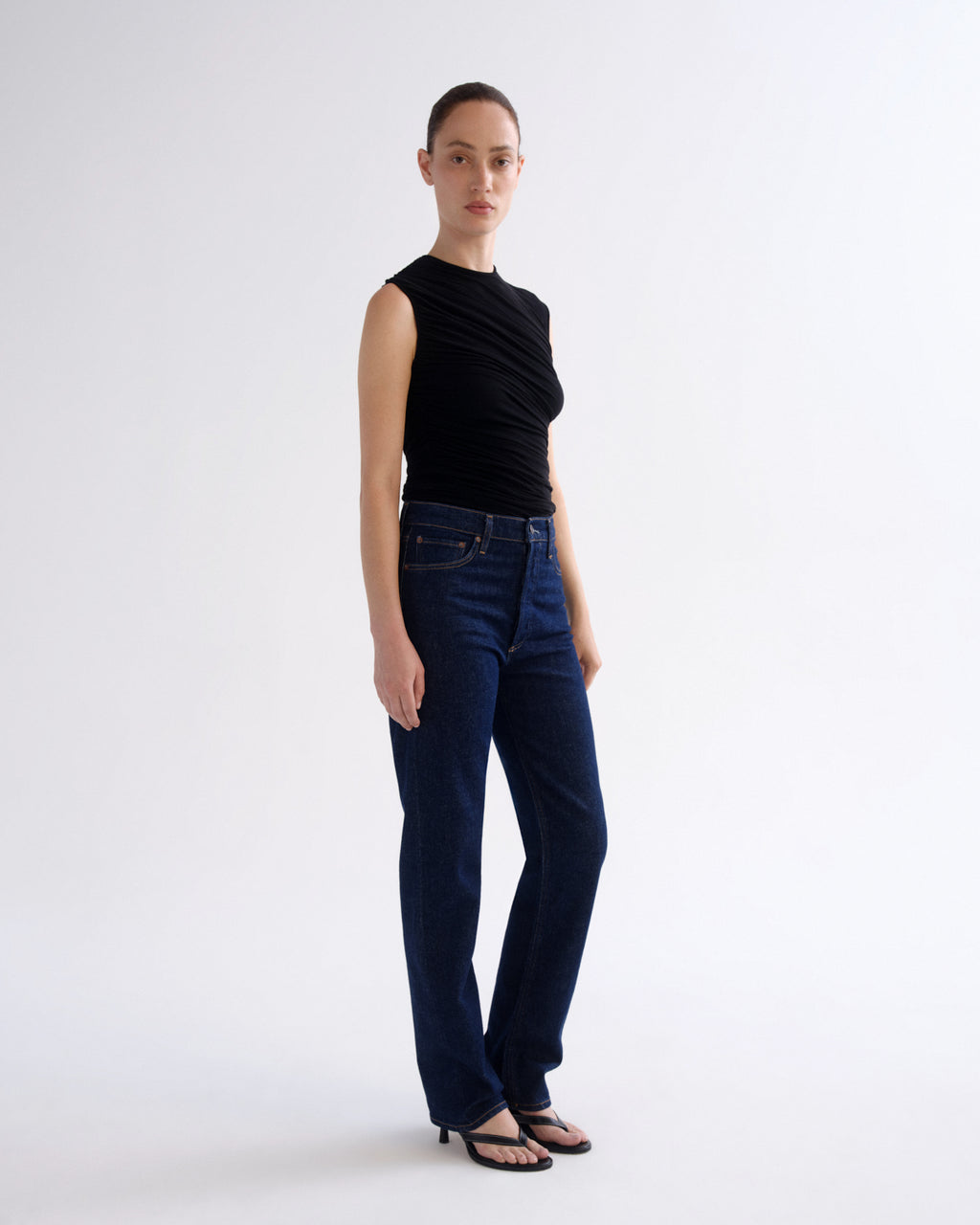 A model stands in a studio wearing a sleeveless black top and dark blue straight-leg jeans with black sandals, against a plain light gray background.