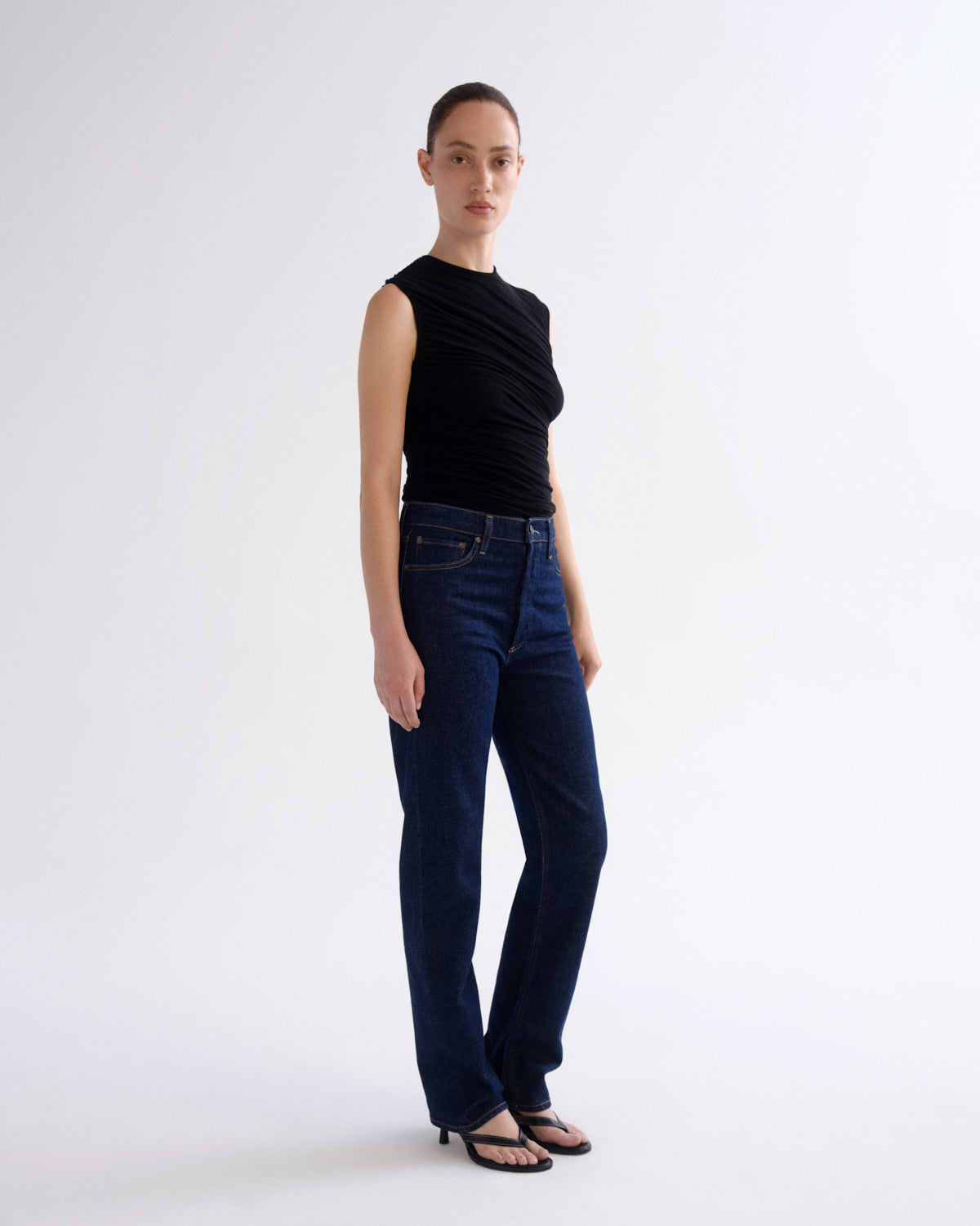 A model stands in a studio wearing a sleeveless black top and dark blue straight-leg jeans with black sandals, against a plain light gray background.