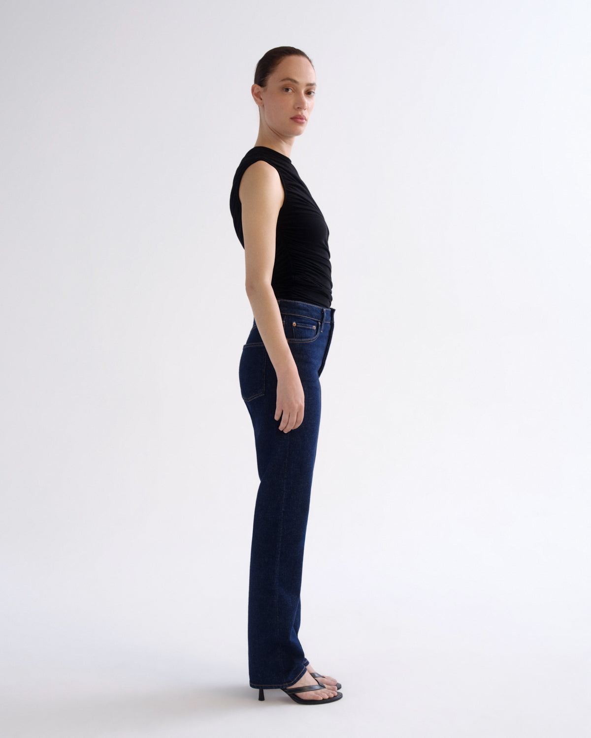 A model standing in profile wearing a black sleeveless top, dark blue high-waisted straight-leg jeans, and black heeled sandals against a plain white studio background.