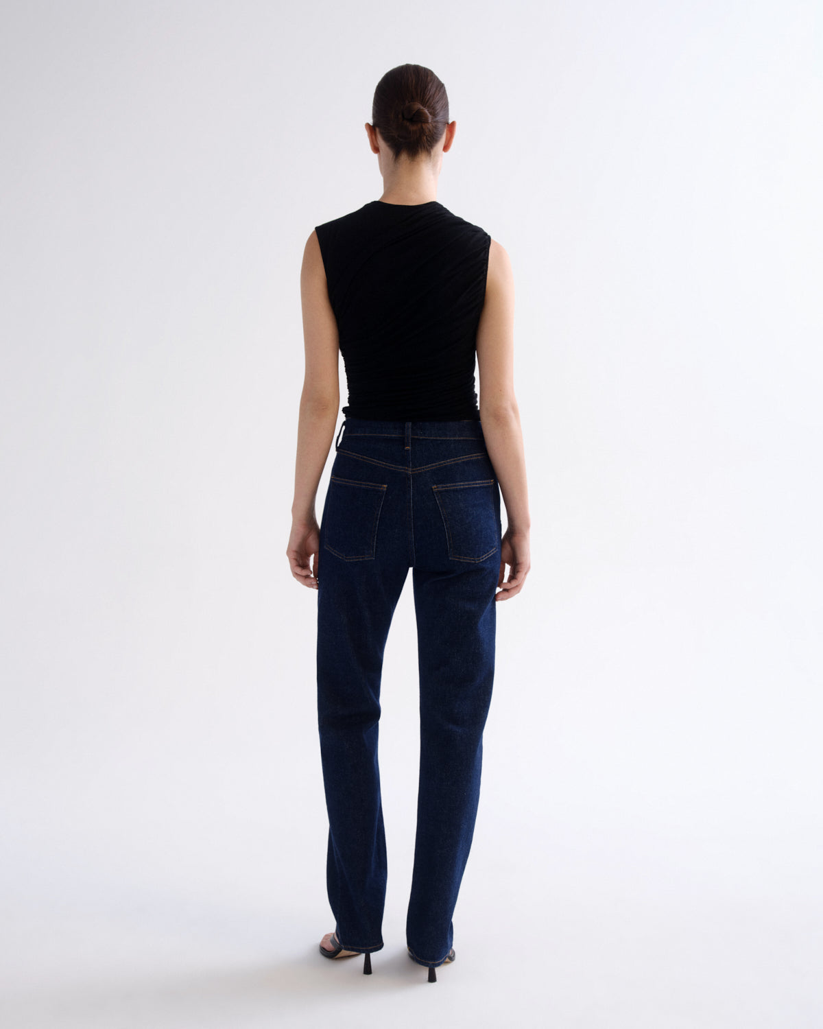 A model viewed from the back wearing a black sleeveless ribbed top and dark blue straight-leg jeans, standing against a plain white studio background.