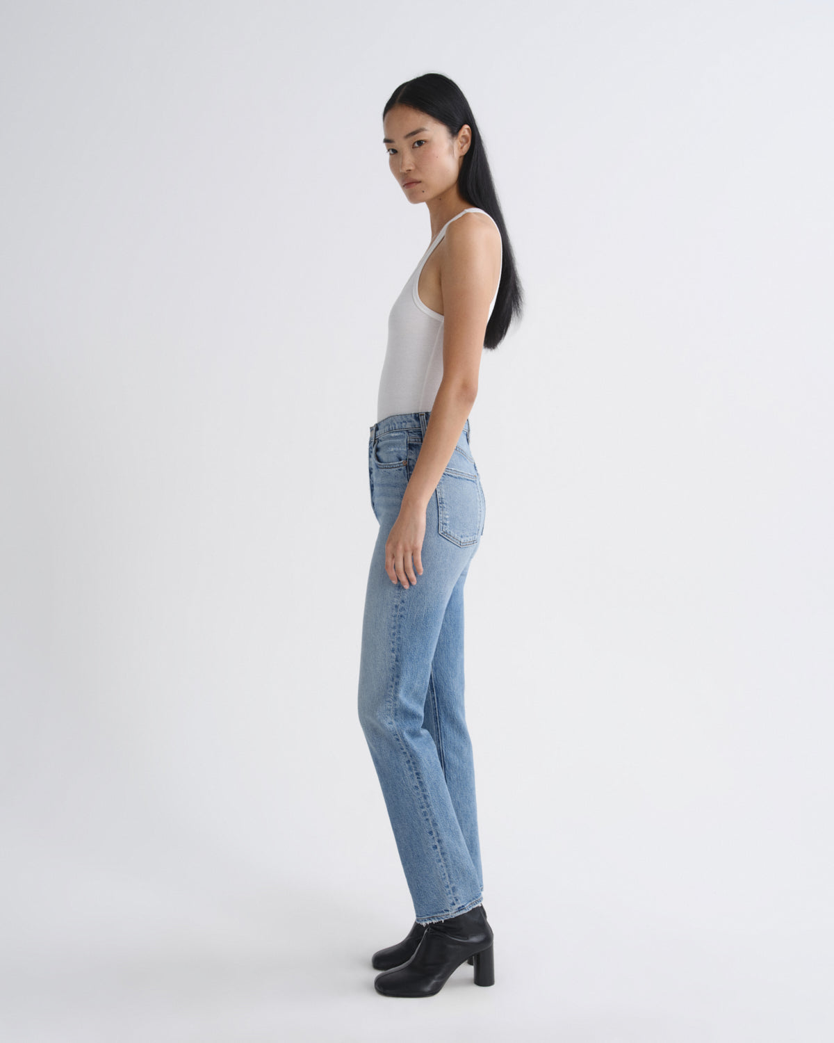 A woman in a white tank top, light-wash blue jeans, and black ankle boots is shown in profile against a plain light gray studio background.