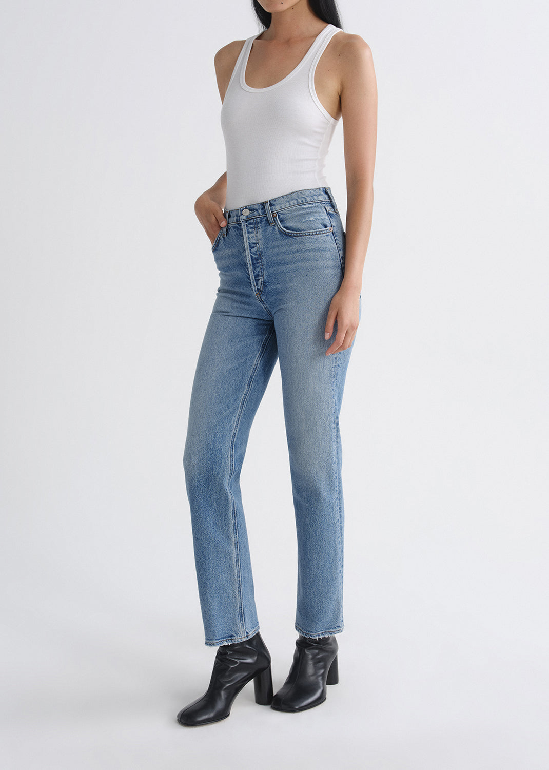 Person wearing light blue jeans and a white tank top on a grey background