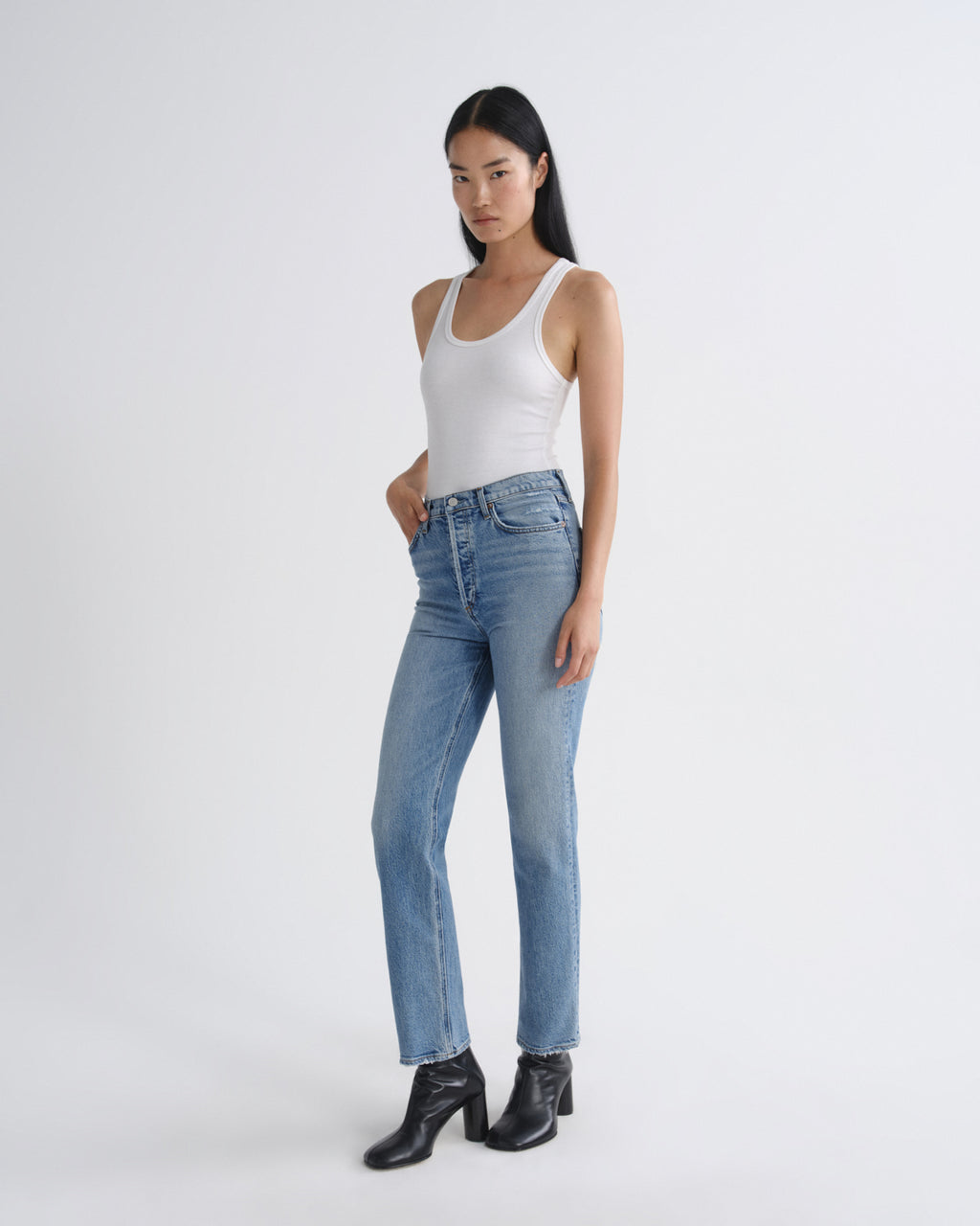 A model wearing light-wash blue straight-leg jeans, a white tank top, and black ankle boots, standing against a plain light gray studio background.