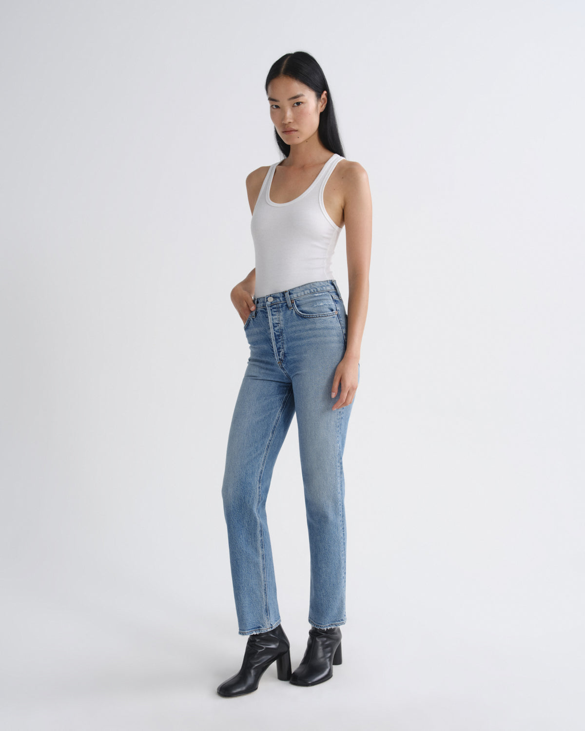 A model wearing light-wash blue straight-leg jeans, a white tank top, and black ankle boots, standing against a plain light gray studio background.