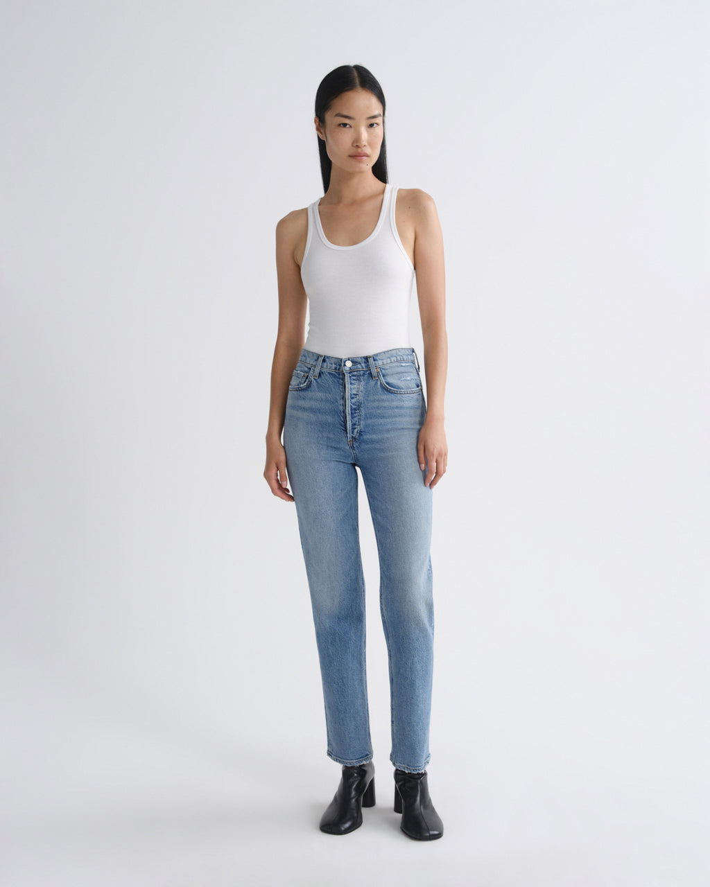 A model stands against a light gray studio background wearing a white ribbed tank top, light-wash blue straight-leg jeans, and black ankle boots.