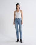 A model stands against a light gray studio background wearing a white ribbed tank top, light-wash blue straight-leg jeans, and black ankle boots.