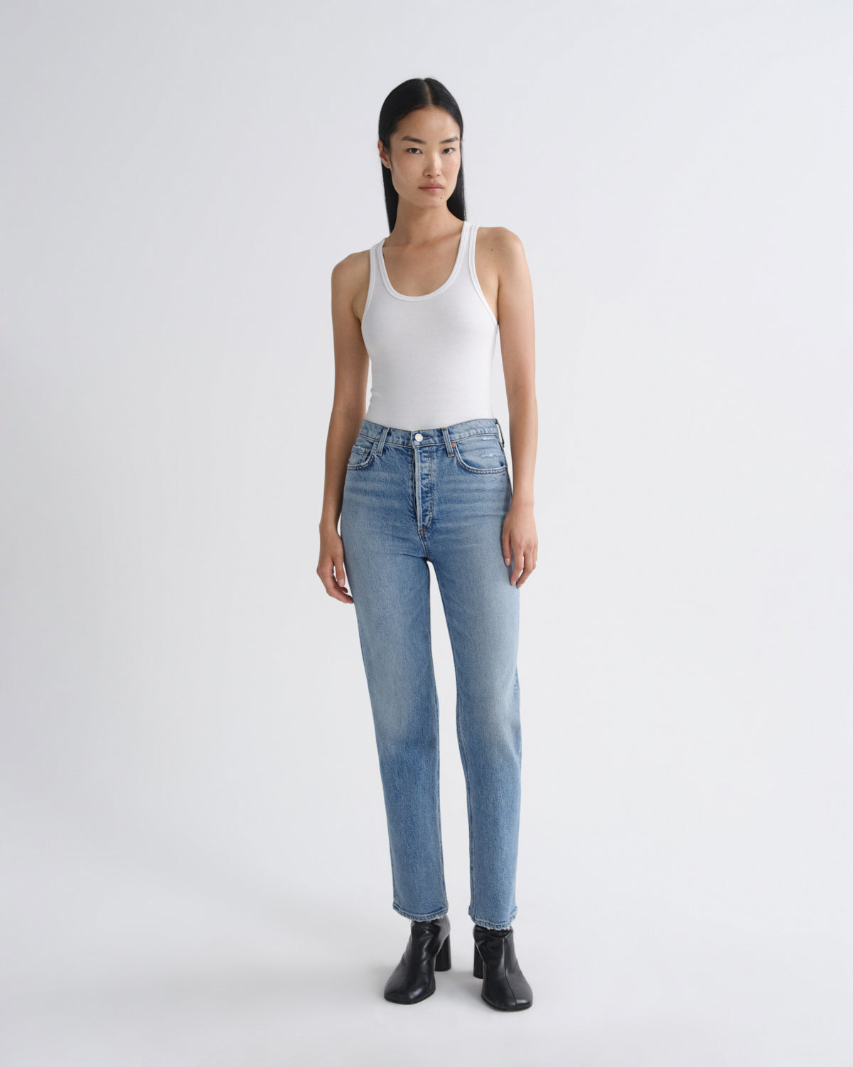A model stands against a light gray studio background wearing a white ribbed tank top, light-wash blue straight-leg jeans, and black ankle boots.