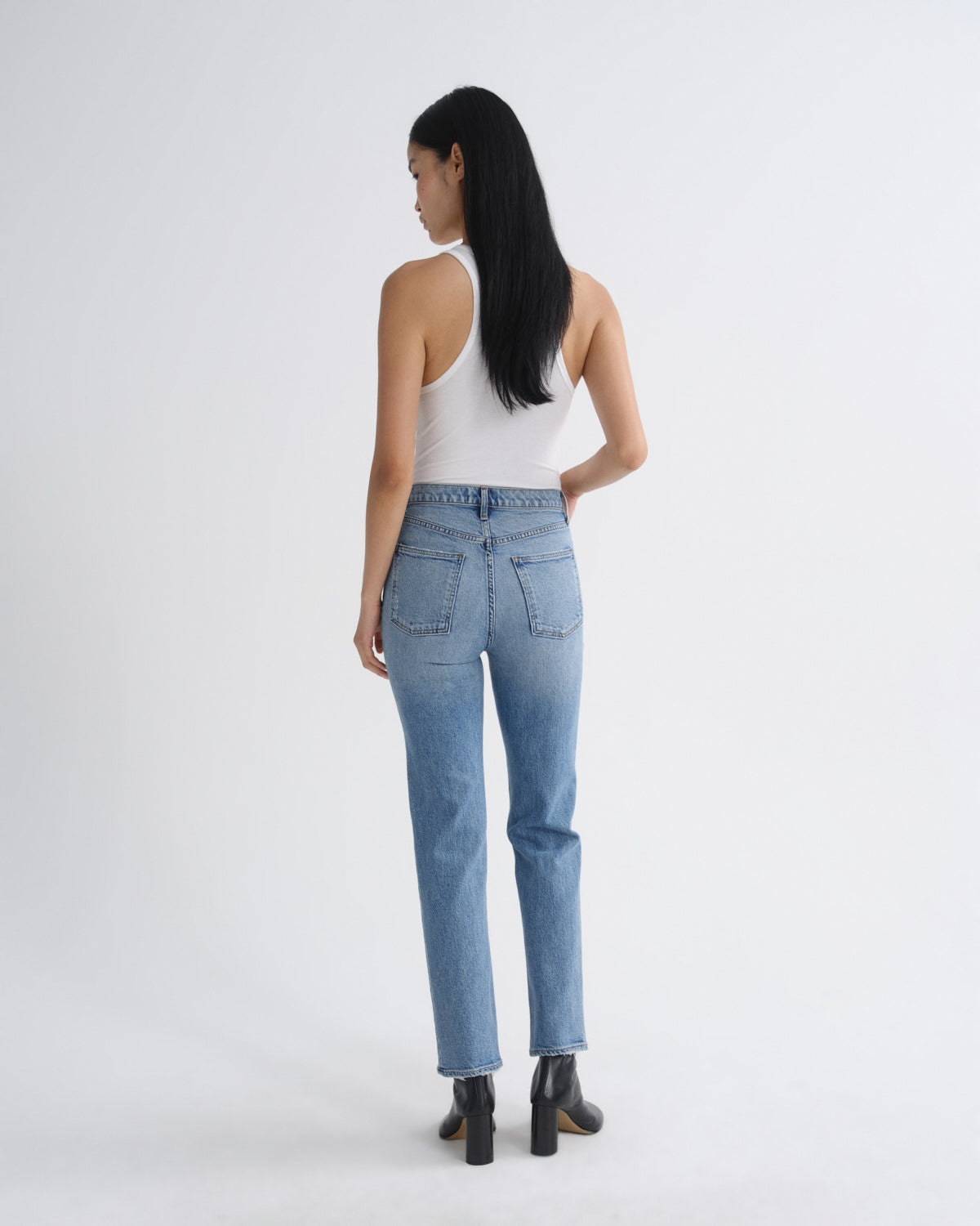 A woman is shown from the back wearing light-wash blue jeans, a white tank top, and black ankle boots against a plain light gray studio background.