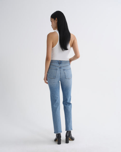 A woman is shown from the back wearing light-wash blue jeans, a white tank top, and black ankle boots against a plain light gray studio background.