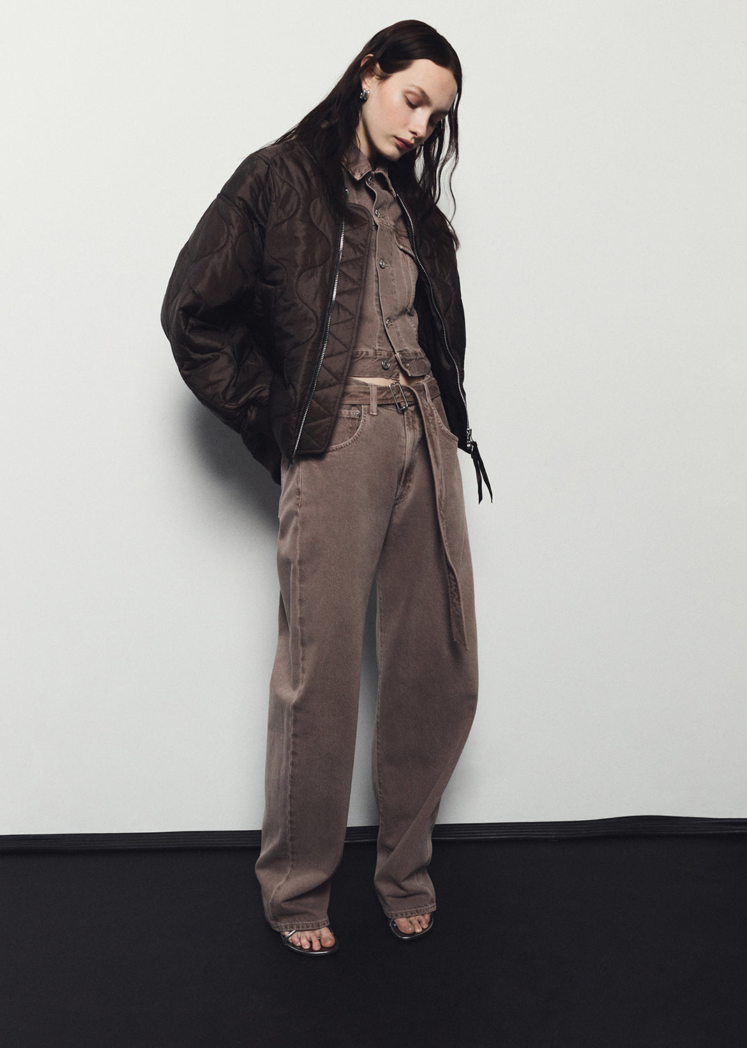 Person wearing a brown jacket and pants against a white wall