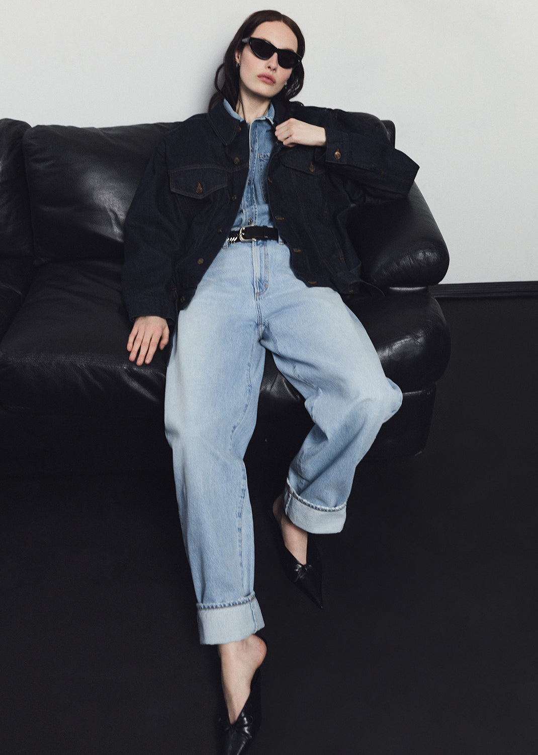 Person wearing a black jacket and light blue jeans sitting on a black leather couch.