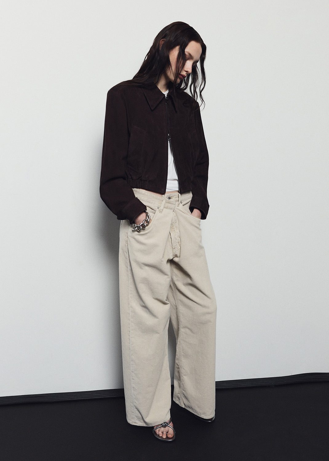 Person wearing a black jacket and beige pants against a white wall