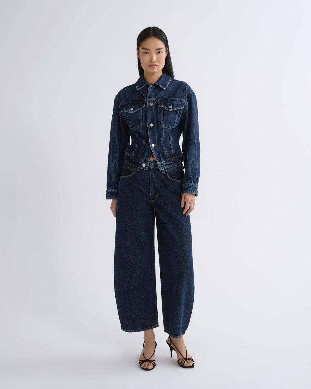 A model stands against a light gray studio background wearing a dark blue denim set consisting of a long-sleeve button-front jacket and matching wide-leg cropped pants, paired with black strappy heels.