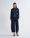 A model stands against a light gray studio background wearing a dark blue denim set consisting of a long-sleeve button-front jacket and matching wide-leg cropped pants, paired with black strappy heels.
