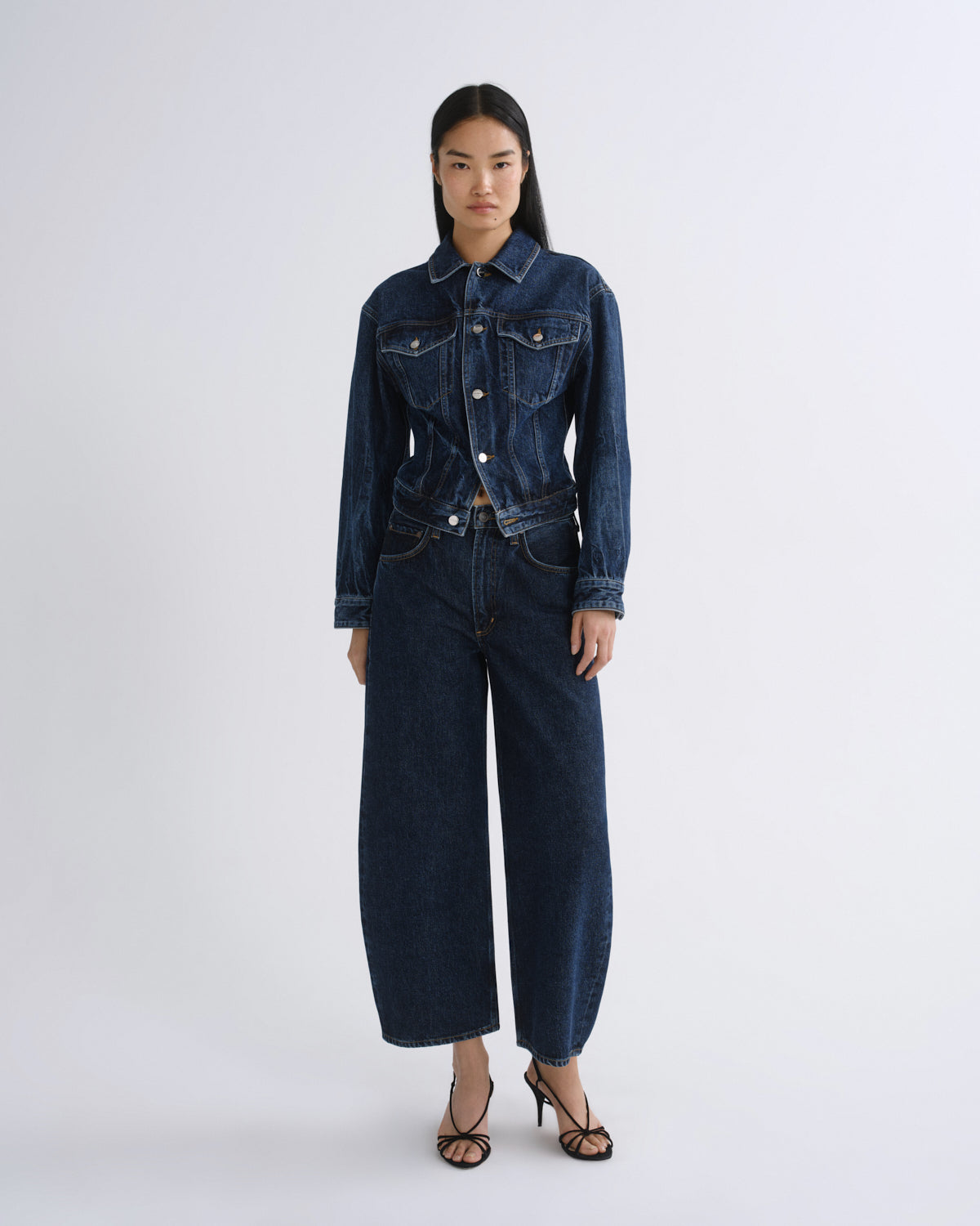 A model stands against a light gray studio background wearing a dark blue denim set consisting of a long-sleeve button-front jacket and matching wide-leg cropped pants, paired with black strappy heels.