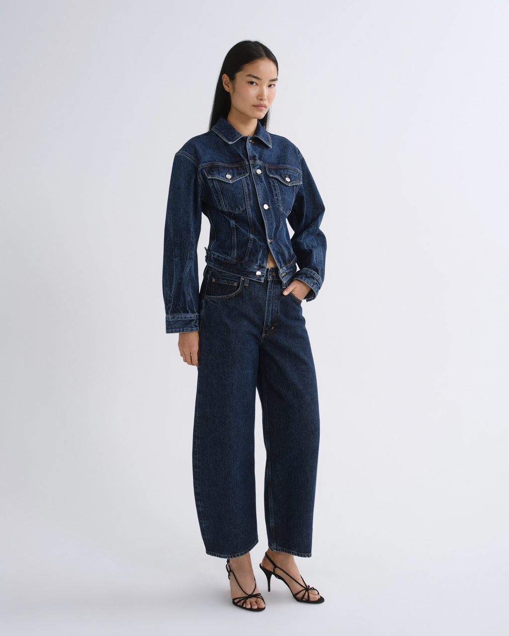 A model stands against a light gray studio background wearing a dark blue denim jacket and matching wide-leg cropped jeans with black strappy heels.