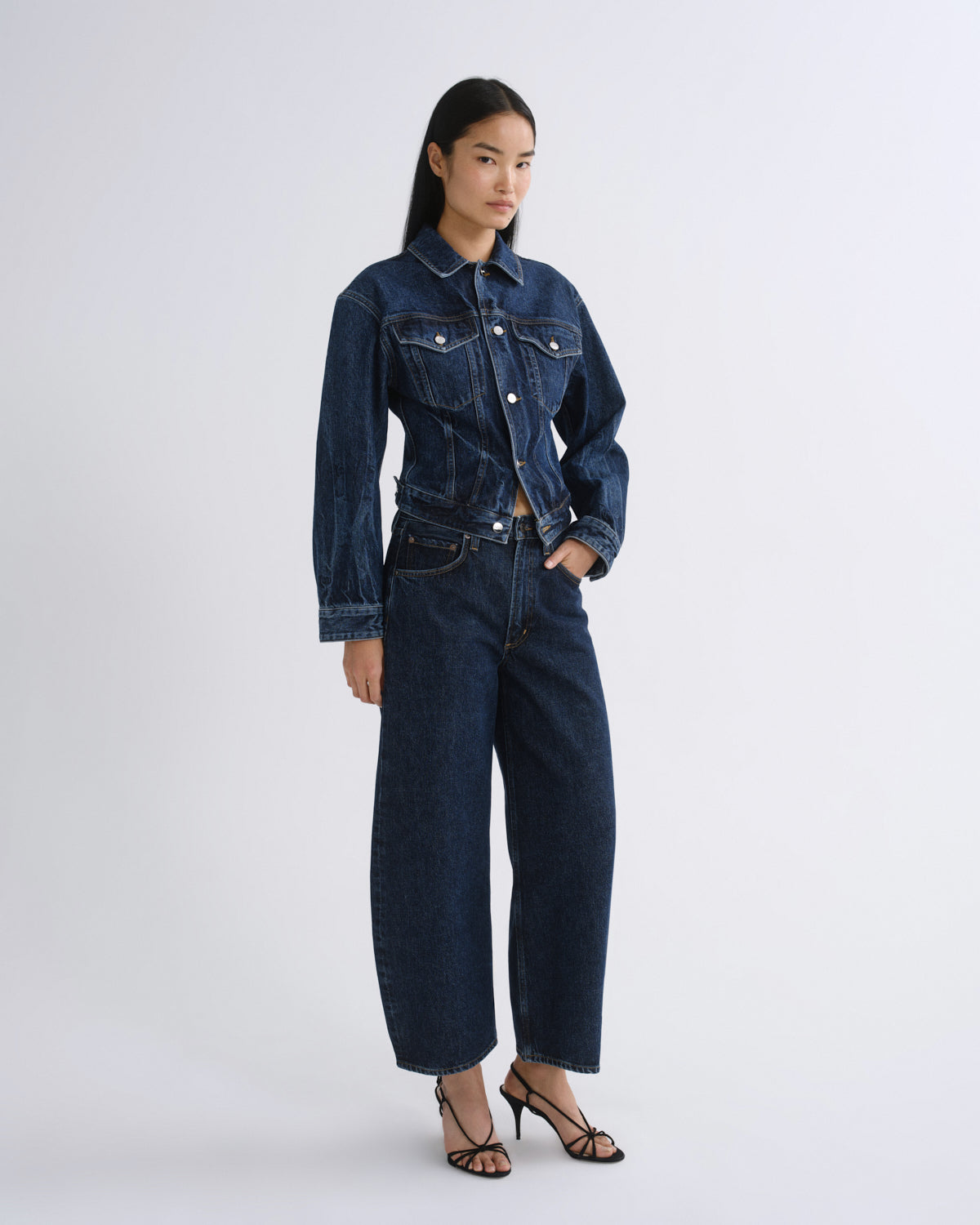 A model stands against a light gray studio background wearing a dark blue denim jacket and matching wide-leg cropped jeans with black strappy heels.
