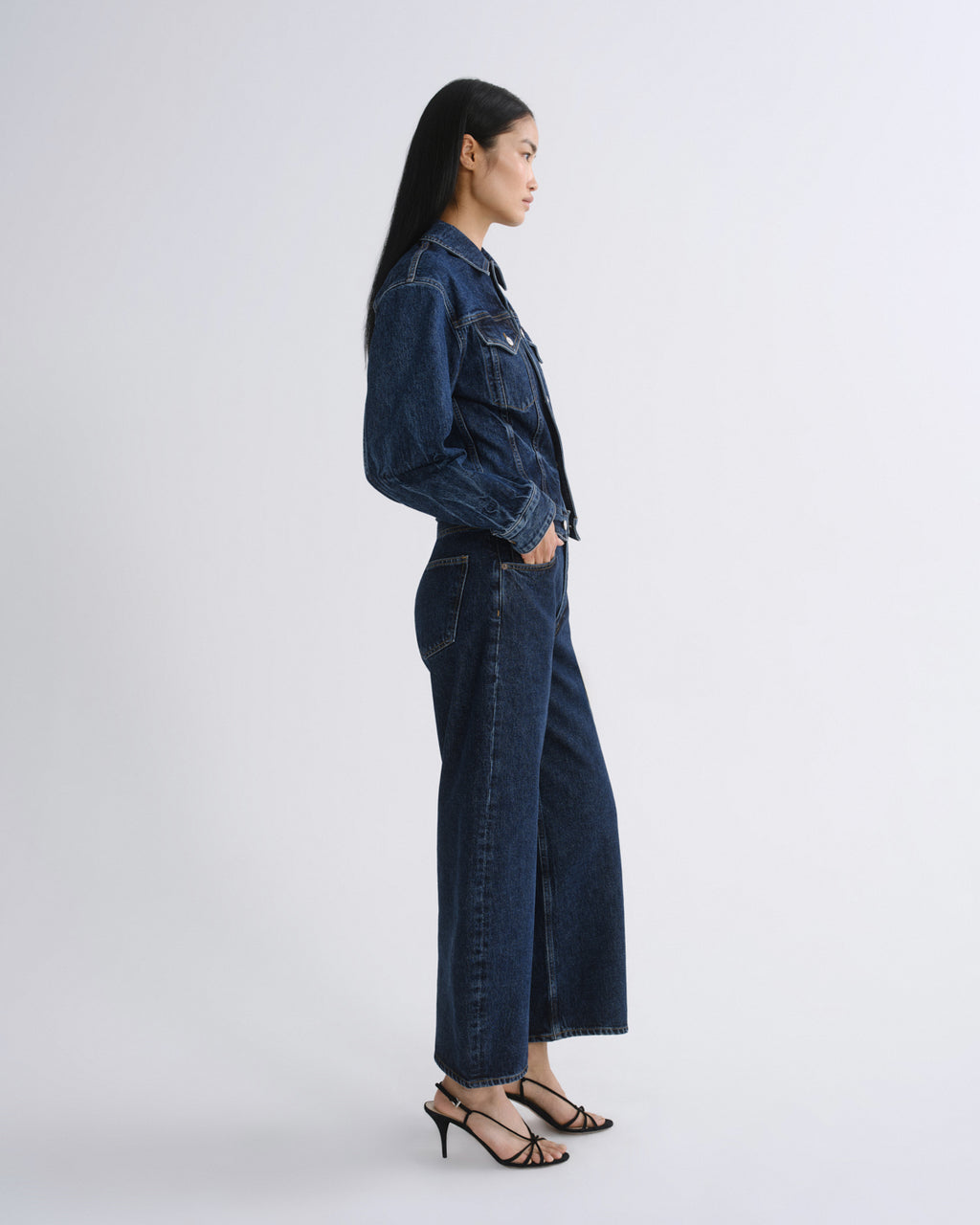 A woman in side profile wearing a dark blue denim jacket and matching wide-leg denim pants, paired with black strappy high-heeled sandals, against a light gray studio background.