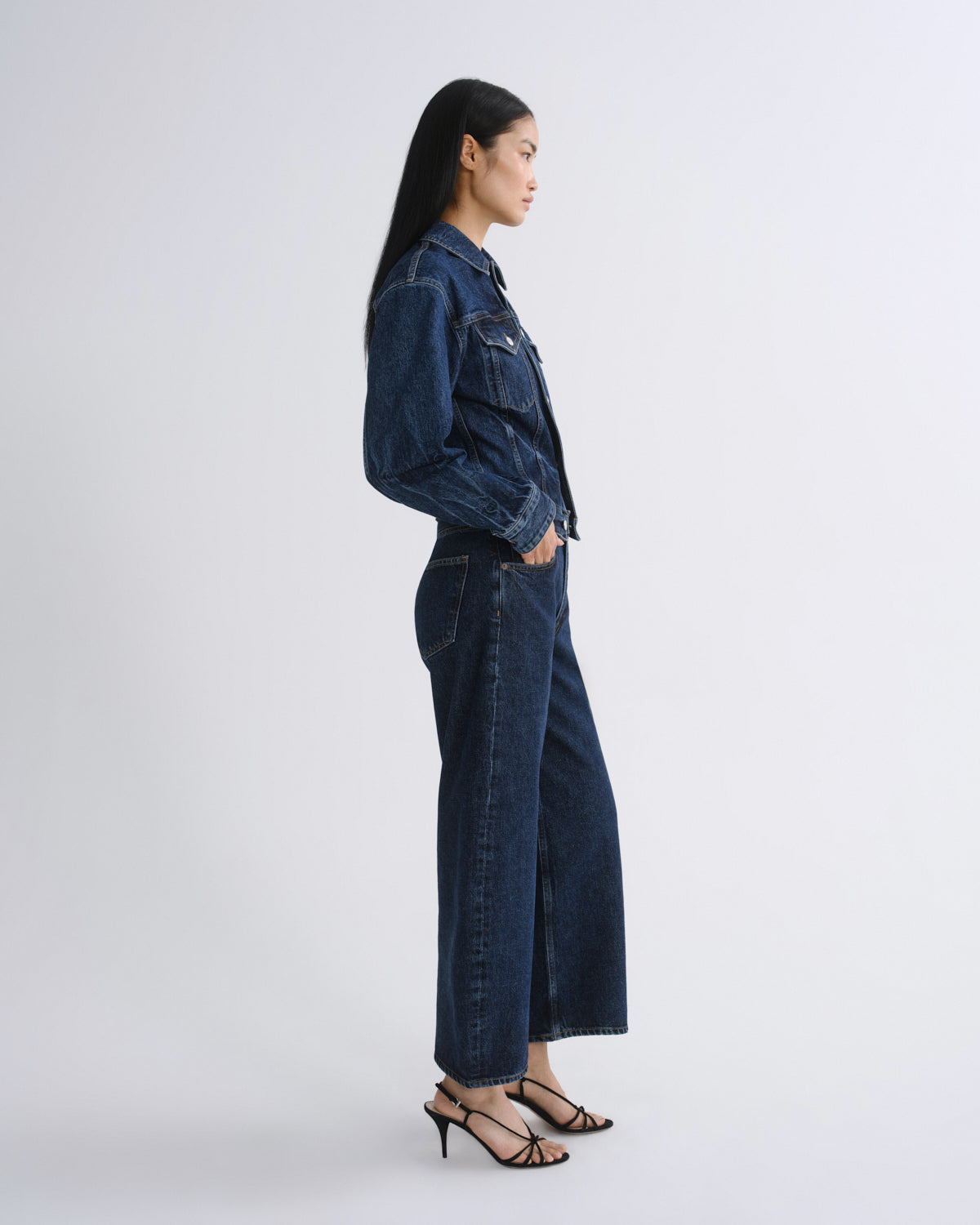 A woman in side profile wearing a dark blue denim jacket and matching wide-leg denim pants, paired with black strappy high-heeled sandals, against a light gray studio background.