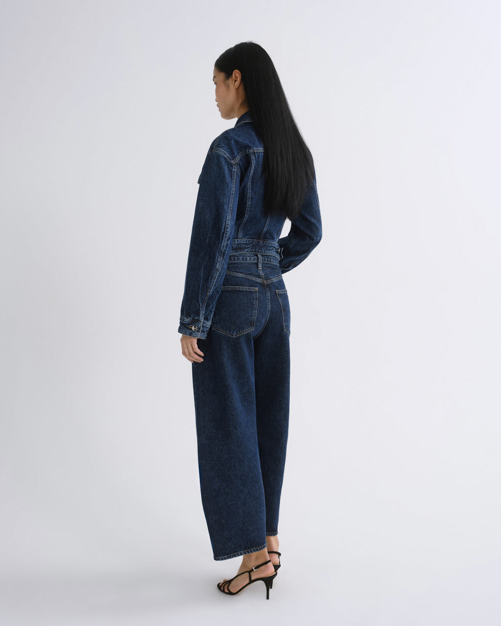A model shown from the back wearing a dark blue denim jumpsuit with wide-leg pants and a belted waist, paired with black strappy high-heeled sandals, against a plain light gray studio background.