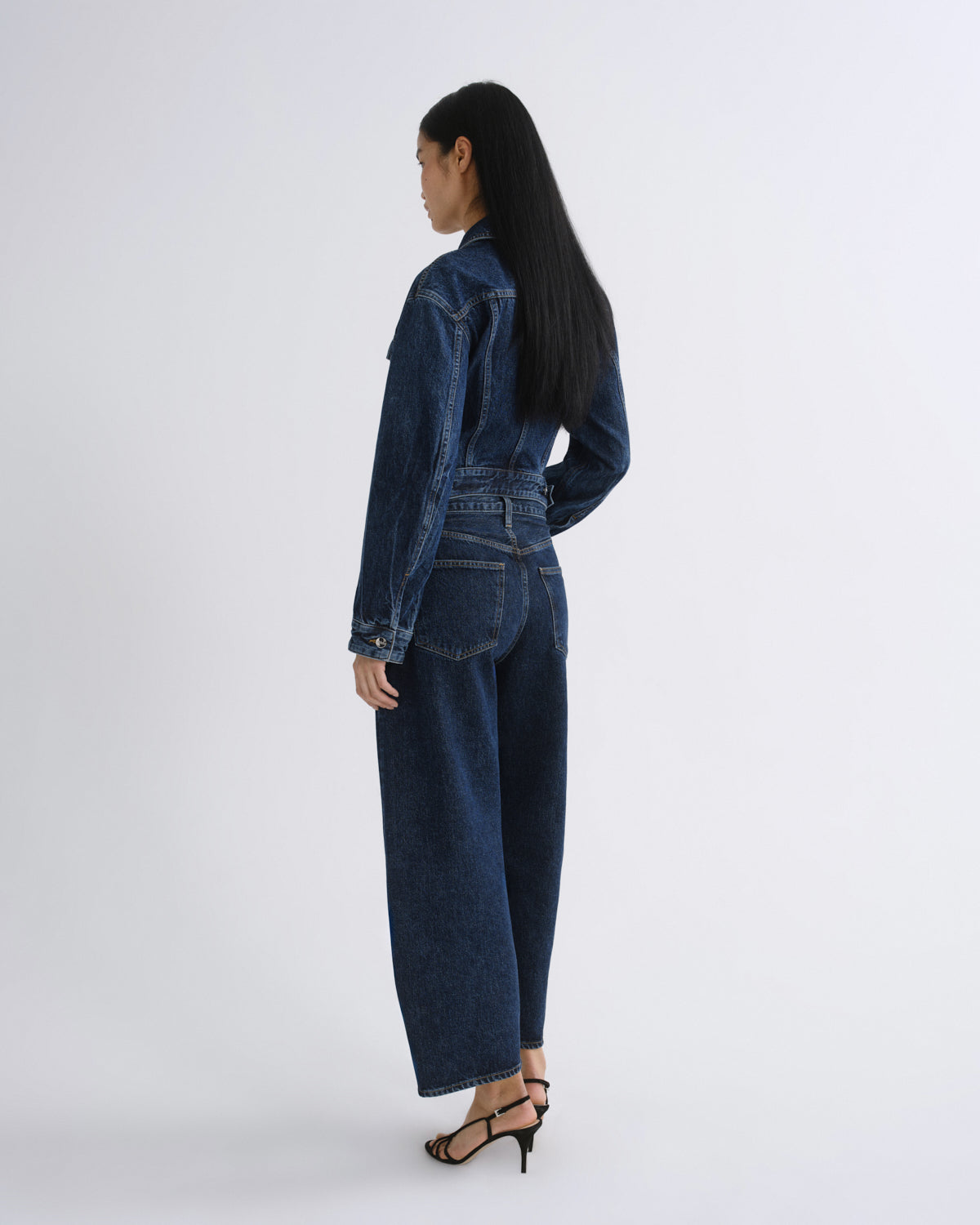 A model shown from the back wearing a dark blue denim jumpsuit with wide-leg pants and a belted waist, paired with black strappy high-heeled sandals, against a plain light gray studio background.