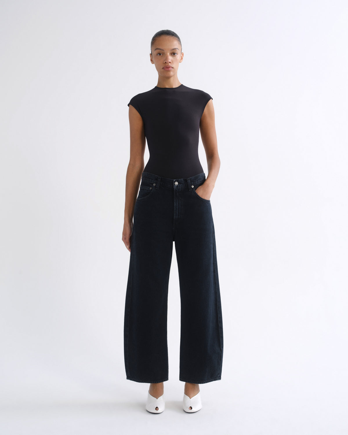 A model stands against a plain white studio background wearing a black sleeveless mock-neck top and black wide-leg cropped jeans, paired with white pointed shoes.