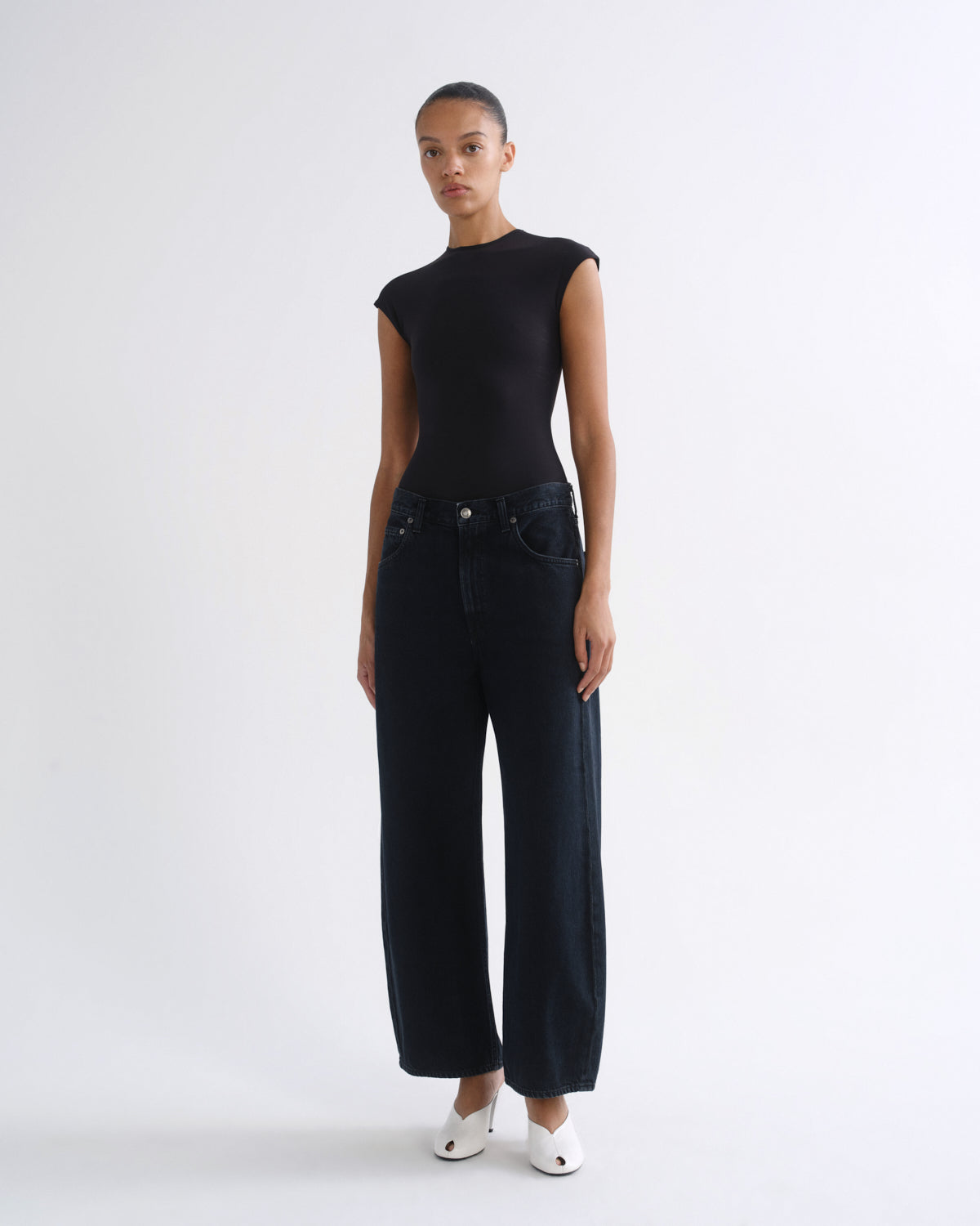 A model stands against a plain white studio background wearing a black short-sleeve top and dark wide-leg jeans, paired with white pointed shoes.