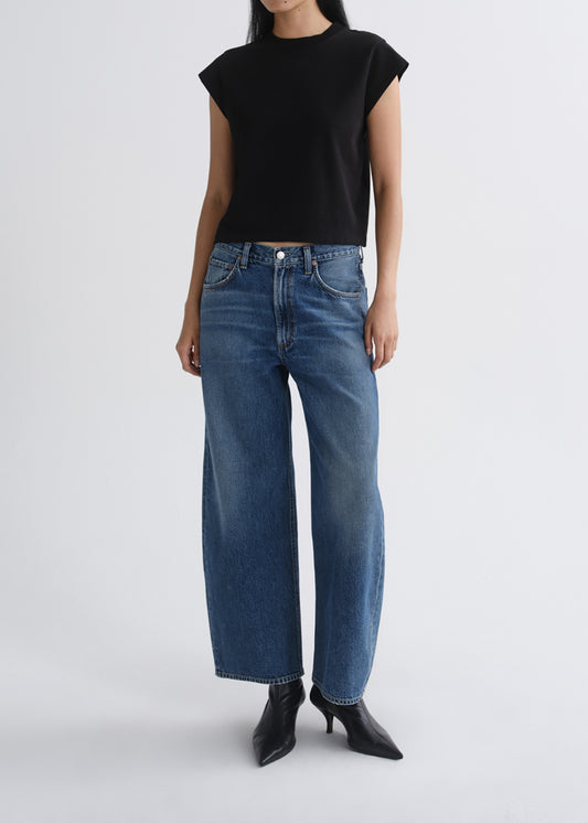 A model wearing a black cropped short-sleeve top and high-waisted, wide-leg blue denim jeans with black pointed-toe ankle boots, photographed against a grey background.