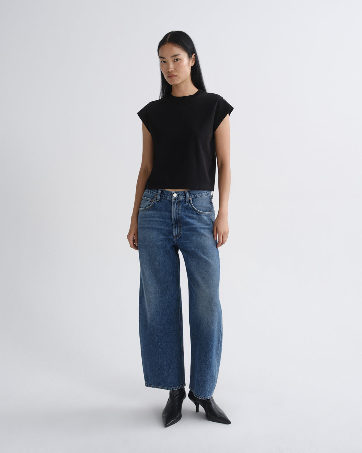 A model stands against a light gray studio background wearing a black short-sleeve top, blue wide-leg jeans, and black pointed ankle boots.
