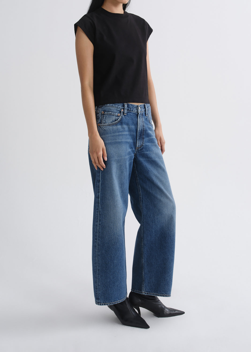 A model wearing a black sleeveless top and wide-leg blue denim jeans with black pointed-toe boots, photographed against a grey background.