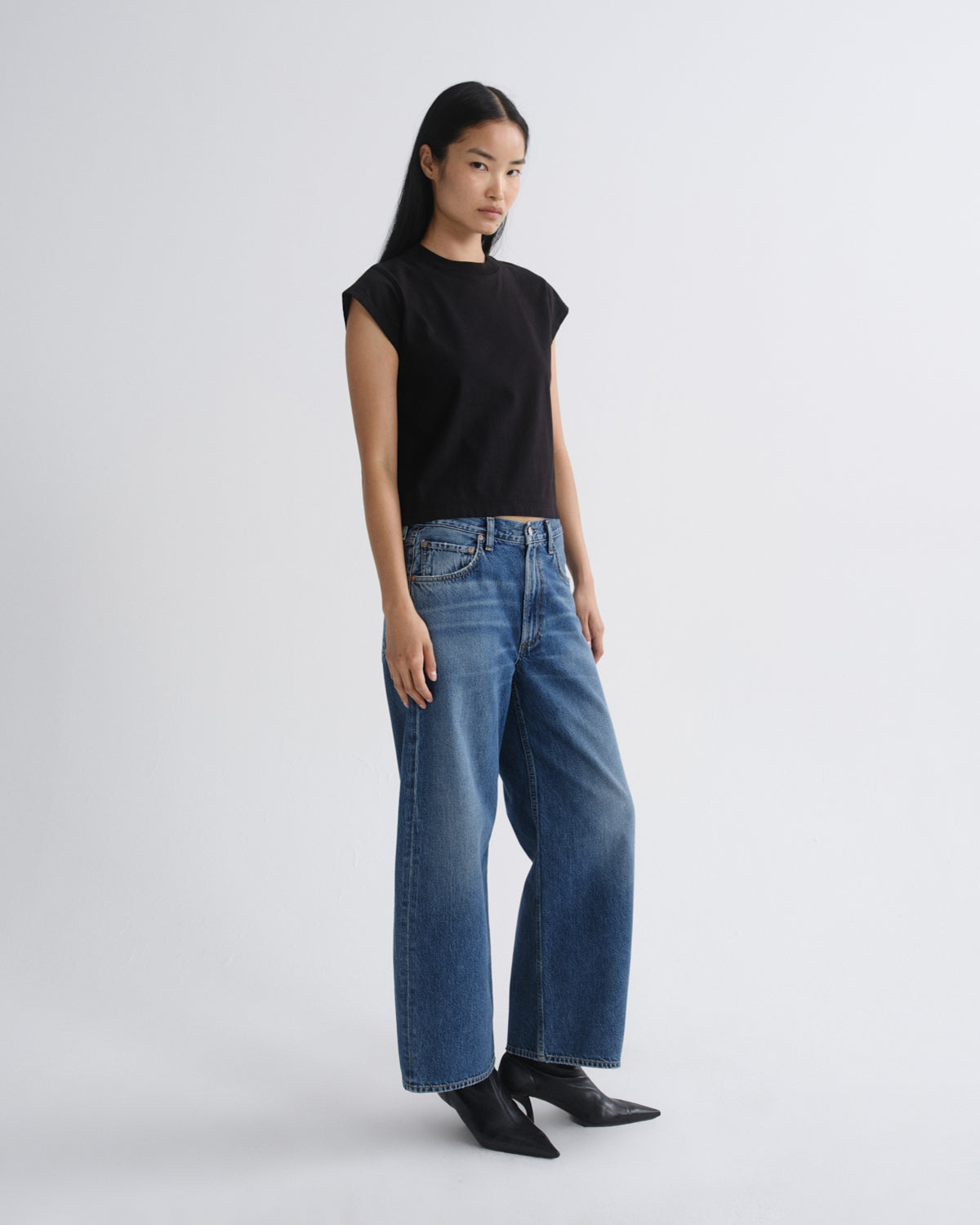 A model stands against a light gray studio background wearing a black short-sleeve top, blue wide-leg jeans, and black pointed ankle boots.