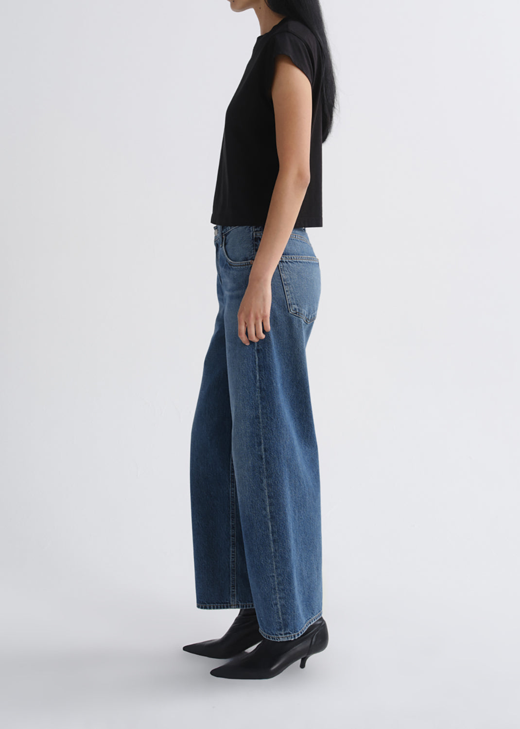 A side-profile studio shot of a model wearing blue wide-leg denim jeans, a black sleeveless top, and black pointed-toe heels against a grey background.